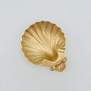 Könnte beinhalten: Eine goldfarbene Metallschale in Form einer Jakobsmuschel. Das Innere weist strahlenförmige Rillen auf, die von der Basis bis zum gewellten Rand verlaufen. Ein kleines, dekoratives scharnierartiges Element mit Muschelmotiv ist unten angebracht.