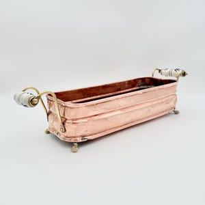 Pode incluir: Uma floreira retangular de cobre com acabamento martelado, apresentando duas alças de latão com detalhes em cerâmica branca e azul. O recipiente é sustentado por quatro pequenos pés de latão, exibindo um brilho metálico e design alongado.
