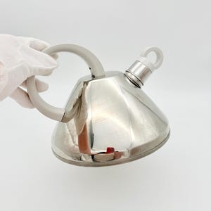 Puede incluir: Una tetera de acero inoxidable pulido con cuerpo de forma cónica. Presenta un asa curva de color gris claro y una tapa de boquilla gris claro a juego con un anillo circular. La superficie reflectante muestra una vista de los alrededores.