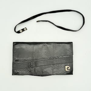 Puede incluir: Bolso de mano de cuero negro con correa desmontable. El bolso de mano presenta un patrón texturizado similar al de un reptil y un logotipo dorado en la parte delantera. La correa tiene dos cierres a presión. El bolso de mano es rectangular y tiene detalles cosidos.