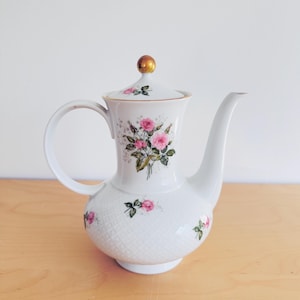 Könnte beinhalten: Weiße Porzellan-Kaffeekanne mit Deckel und Henkel. Verziert mit rosa Rosen und grünen Blättern. Der Deckel hat einen goldenen Knopf. Die Kanne hat ein Rautenmuster auf der unteren Hälfte.