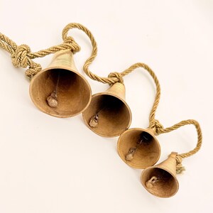 May include: Four brass bells of varying sizes are suspended from a twisted rope. The bells have a warm, golden hue and a small clapper inside. The rope is a natural, beige color, and the bells are arranged diagonally.