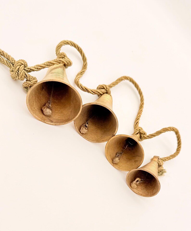 May include: Four brass bells of varying sizes are suspended from a twisted rope. The bells have a warm, golden hue and a small clapper inside. The rope is a natural, beige color, and the bells are arranged diagonally.