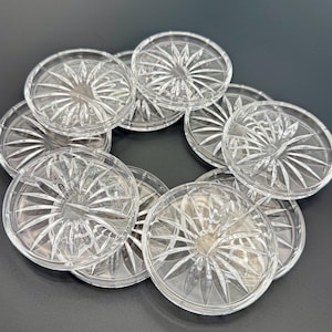 Op de afbeelding: Een verzameling heldere, ronde glazen onderzetters met een sterrenpatroon. De onderzetters zijn in een cirkelvorm gerangschikt, waarbij hun ingewikkelde, geëtste details worden getoond. Het glas heeft een reflecterende kwaliteit, waardoor het licht wordt opgevangen.