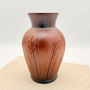 Może przedstawiać: Brązowy ceramiczny wazon z ciemnobrązową obwódką i rzeźbionym wzorem liści. Wazon ma około 15 cm wysokości i stoi na jasnej powierzchni. Wazon ma lekko rozszerzoną górę i zaokrąglony korpus.