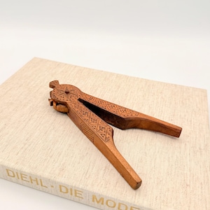 May include: A wooden nutcracker with intricate geometric carvings. The nutcracker is open, revealing the interior mechanism. The tool is resting on a book with the text "DIEHL - DIE MODERNEN" in gold lettering.