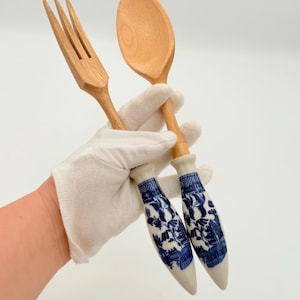 Puede incluir: Juego de tenedor y cuchara de madera. El tenedor y la cuchara son de madera clara. Los mangos son blancos con un diseño floral azul. El conjunto lo sostiene una mano enguantada.