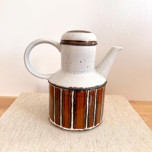 Peut inclure: Une cafetière en céramique avec un corps blanc moucheté et une base à rayures marron. Le couvercle et le bord sont marron. La poignée et le bec sont blancs. La cafetière est posée sur une pile de livres.