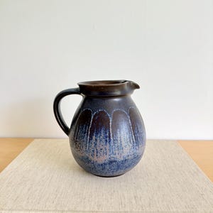May include: A ceramic pitcher with a dark blue glaze and a brown rim and handle. The pitcher has a rounded body with a spout and a curved handle. The pitcher is sitting on a book with gold lettering. The pitcher is approximately 7 inches tall.