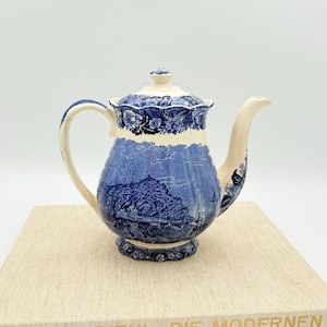May include: A vintage blue and white ceramic teapot with a lid, handle, and spout. The teapot features a landscape design and floral accents. It is placed on a light-colored surface.