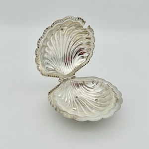 May include: A silver-colored, open clam shell-shaped trinket box. The box is hinged and has a detailed, ridged design on both halves. The edges are scalloped and adorned with small, round details. The interior reflects the light.