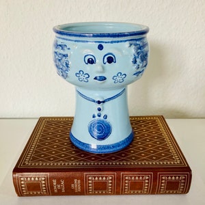 Pode incluir: Um vaso de cerâmica azul claro com um design de rosto, com detalhes azuis e brancos. O vaso tem um topo redondo e um pescoço com um design de pingente. É colocado em um livro marrom com letras douradas.