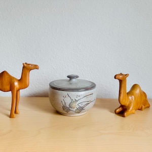 Könnte beinhalten: Zwei Holz-Kamel-Figuren und ein Keramiktopf mit Deckel. Die Kamele sind aus hellem Holz geschnitzt. Der Topf hat ein gesprenkeltes Design in Braun und Grau. Ein Kamel steht, das andere liegt.