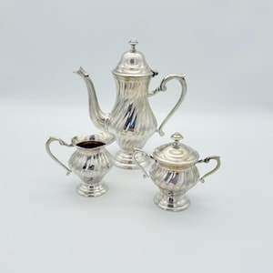 Może przedstawiać: Srebrny zestaw do herbaty z dzbankiem do kawy, śmietanką i cukiernicą. Zestaw ma żebrowany wzór i jest prezentowany na białym tle. Dzbanek do kawy ma pokrywkę i zakrzywiony uchwyt. Śmietanka i cukiernica również mają uchwyty.