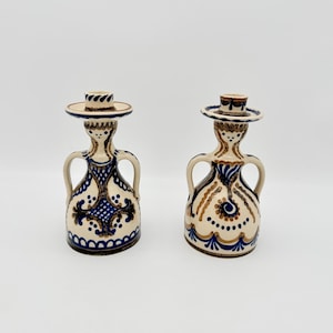 May include: Two ceramic candle holders with a unique design. The candle holders are off-white with blue and brown decorative patterns. Each has a small hat-like top and two handles. The candle holders are approximately 6 inches tall.