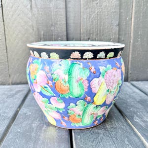 Può includere: Un vaso in ceramica rotondo con uno sfondo blu vivace decorato con illustrazioni colorate di vari frutti e verdure. Il bordo è nero con un motivo ripetitivo di piccoli fiori stilizzati in tonalità bianche e pastello delicate.