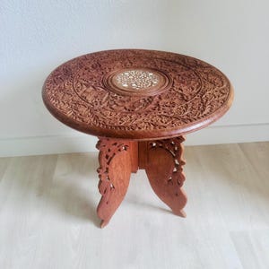 Può includere: Un tavolino rotondo in legno caratterizzato da intricati intagli floreali sulla superficie e sulle gambe. Il centro del piano del tavolo presenta un intarsio circolare con un motivo bianco dettagliato. Il tavolo ha tre gambe intagliate.