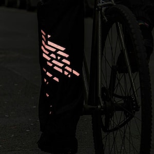 Puede incluir: Una persona montando en bicicleta por la noche con tiras reflectantes en sus pantalones. Las tiras reflectantes son un patrón geométrico de color rosa claro.
