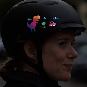 Könnte beinhalten: Eine Person trägt einen schwarzen Fahrradhelm mit einem Regenbogen-Dinosaurier, einem grünen Baum und einem rosa Autoaufkleber auf der Vorderseite.