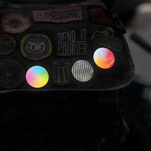 Könnte beinhalten: Ein schwarzer Rucksack mit einer Vielzahl von bunten Patches und Aufklebern, darunter ein regenbogenfarbener reflektierender Aufkleber, ein weiß-schwarz gestreifter reflektierender Aufkleber und ein rot-orange-gelber reflektierender Aufkleber. Der Rucksack hat auch einen Patch mit der Aufschrift "Send It Courier".