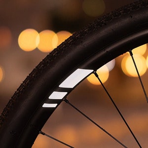 Könnte beinhalten: Nahaufnahme eines schwarzen Fahrradrads mit reflektierenden weißen Streifen. Das Rad hat schwarze Speichen und einen schwarzen Reifen. Der Hintergrund ist verschwommen und unscharf.