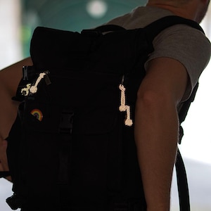 May include: A black rucksack with reflective zip pulls and a rainbow patch. The rucksack has multiple compartments and straps. The reflective zip pulls are shaped like scissors and a bone. The rucksack is being carried.