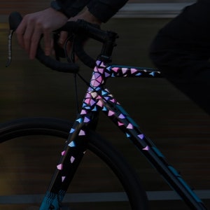 May include: A black bicycle frame with a colourful geometric pattern. The pattern is made up of pink, blue, and purple triangles. The bicycle is being ridden at night.