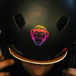 Könnte beinhalten: Ein schwarzer Helm mit einem regenbogenfarbenen Affen-Motiv auf der Vorderseite. Der Helm hat einen braunen Rand.