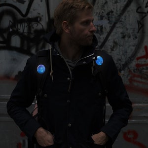 Könnte beinhalten: Eine Person trägt eine schwarze Jacke mit zwei leuchtenden, runden, blau-violetten Anstecknadeln, die an der Jacke und den Rucksackgurten befestigt sind. Die Anstecknadeln haben ein galaxieartiges Design. Der Hintergrund ist eine Graffiti-Wand.