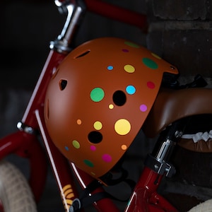 Peut inclure: Casque de vélo marron orné de pois multicolores. Le casque est posé sur une selle et un cadre de vélo rouges. Il comporte des trous d'aération pour une meilleure circulation de l'air.
