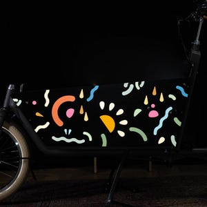 May include: A black cargo bike with a colorful abstract design on the side. The design features various shapes and patterns in shades of pink, orange, yellow, blue, and green.