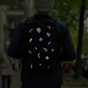 Könnte beinhalten: Schwarzer Rucksack mit reflektierenden, bunten geometrischen Formen. Die Formen umfassen Wirbel, Dreiecke und Kreuze. Der Rucksack wird von einer Person in einer dunklen Jacke und einer Mütze getragen.
