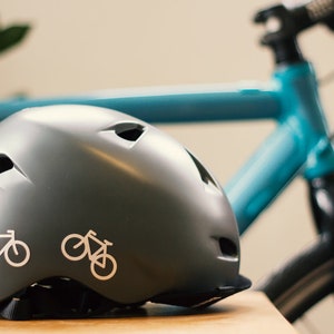 Op de afbeelding: Een grijze fietshelm met twee witte fiets stickers aan de zijkant. De helm ligt op een houten oppervlak met een blauwe fiets op de achtergrond.