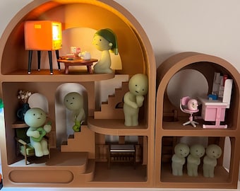 May include: A multi-level tan dollhouse with arched openings containing several pale green humanoid figurines. The interior features miniature furniture including a television, table, chairs, and a desk. A street lamp with a glowing green shade stands to the left.