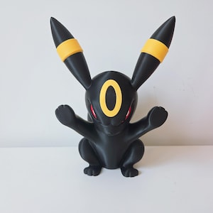 May include: A black figurine of the Pokemon character Umbreon with long ears featuring yellow bands, a yellow oval marking on the forehead, and red eyes. The creature is in a seated position with both front paws raised in a forward-facing pose.