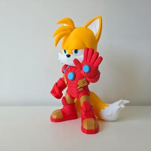 May include: A figurine of Tails from Sonic the Hedgehog, dressed in a red and gold Iron Man suit. The figure has yellow fur, a white muzzle, and blue eyes. The figure is standing with one hand raised.