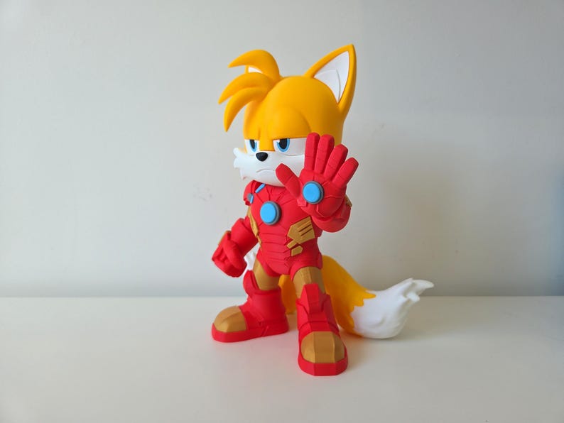 May include: A figurine of Tails from Sonic the Hedgehog, dressed in a red and gold Iron Man suit. The figure has yellow fur, a white muzzle, and blue eyes. The figure is standing with one hand raised.