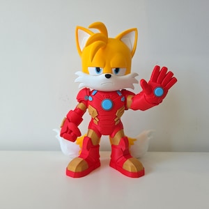 May include: A yellow and white Tails the Fox figurine wearing a red and gold Iron Man suit. The figurine has a stern expression and is posed with one hand raised. The figure is on a white surface.