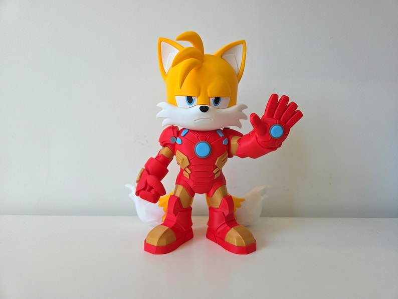 May include: A yellow and white Tails the Fox figurine wearing a red and gold Iron Man suit. The figurine has a stern expression and is posed with one hand raised. The figure is on a white surface.