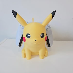 May include: A yellow Pikachu character figure serving as a controller stand. The figure features long ears with black tips, red circular cheeks, and black eyes. A white and black gaming controller is positioned behind the figure's back.