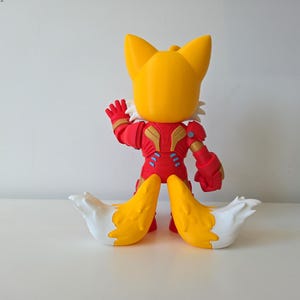 May include: A yellow and red figurine of a fox-like character with large ears and two tails. The figure is wearing a red and gold suit with blue accents. The tails are yellow with white tips. The figure is posed with one arm raised.