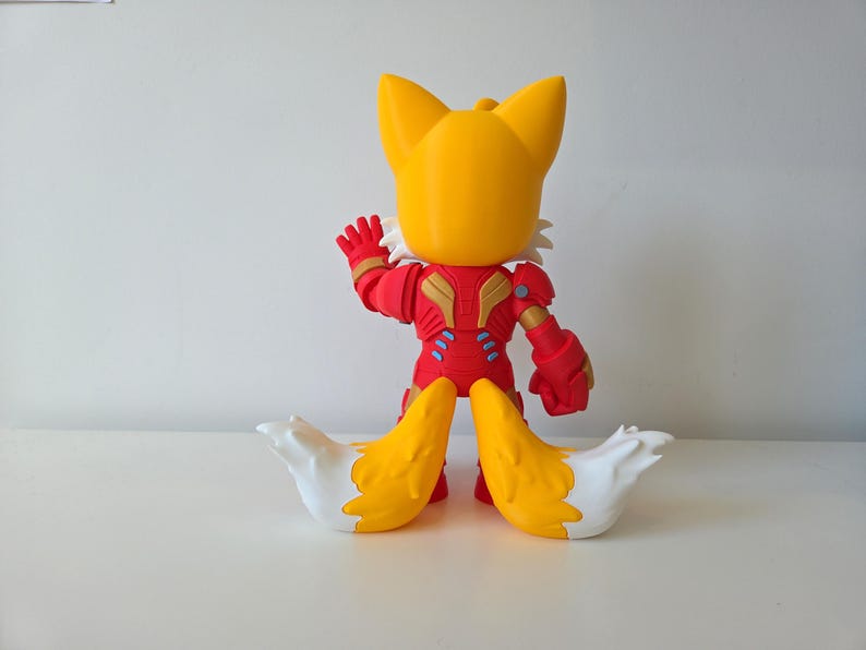 May include: A yellow and red figurine of a fox-like character with large ears and two tails. The figure is wearing a red and gold suit with blue accents. The tails are yellow with white tips. The figure is posed with one arm raised.