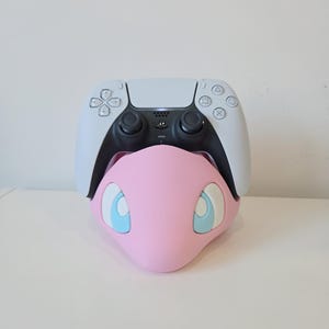 May include: A white and black wireless game controller resting on a light pink stand shaped like a creature head. The stand features large, stylised light blue and white eyes on the front, providing a stable base for the gaming peripheral.