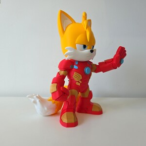 May include: A figurine of Tails from Sonic the Hedgehog, wearing a red and gold Iron Man suit. The figure has a yellow head with two ears, a white muzzle, and a white and yellow tail. The Iron Man suit has a blue circle on the chest.