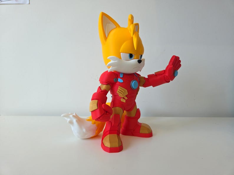 May include: A figurine of Tails from Sonic the Hedgehog, wearing a red and gold Iron Man suit. The figure has a yellow head with two ears, a white muzzle, and a white and yellow tail. The Iron Man suit has a blue circle on the chest.