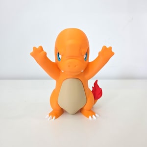 Peut inclure: Figurine orange de Charmander avec les bras levés. Le personnage a un ventre beige, des yeux bleus et une flamme rouge sur la queue. Des griffes blanches sont visibles sur ses pieds.