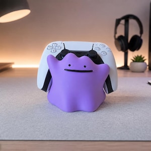 May include: A purple, blob-shaped controller stand featuring a simple face with two black dots for eyes and a thin black line for a mouth. The stand is holding a white and black wireless game controller securely in its center.