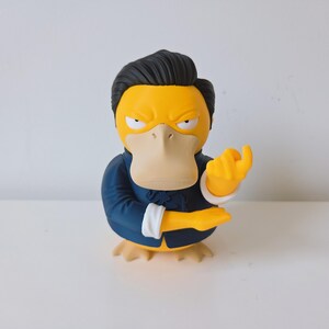 May include: A yellow duck-like character figurine with black hair, wearing a dark blue blazer with white cuffs. The character has a stern facial expression with narrowed eyes and a beige beak, posed with one hand gesturing forward in a beckoning motion.
