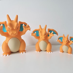 May include: Three orange dragon-like figures of varying sizes, each featuring a cream-coloured belly, teal-coloured inner wings, and white claws. The figures are standing in a row, with the largest on the left and the smallest on the right.