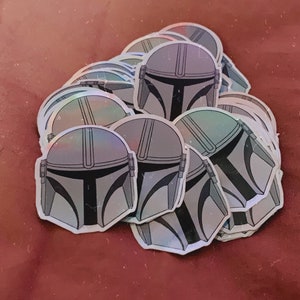 May include: A pile of holographic stickers featuring the Mandalorian helmet design. The stickers have a silver and gray color scheme with black accents. The stickers are on a maroon background.